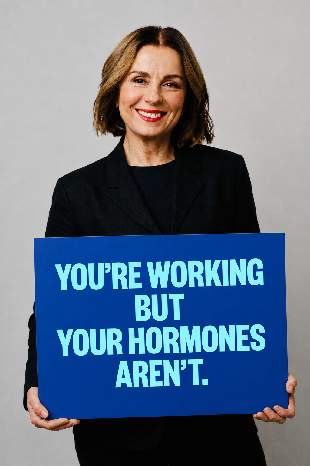 Woman in a black blazer holding a blue sign reading: You're working but your hormones aren't.